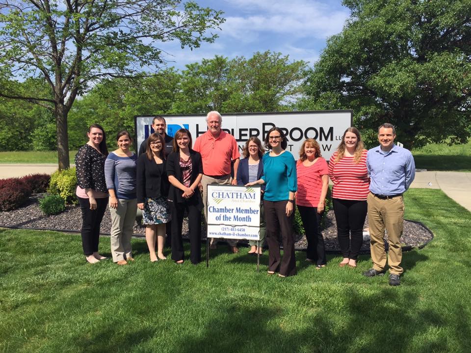 Group of 7 women and 3 men standing outside in front of a large sign for HeplerBroom. In front of the group is a small sign for Chatham Chamber Member of the Month. In the background are trees and a piece of clear blue sky.'