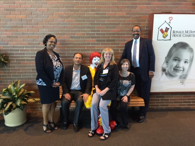 3 women and 2 men sitting/standing around bench on which Ronald McDonald is sitting. In background is a brick wall and a part of a poster for Ronald McDonald House Charity with with a little girl's face on it.