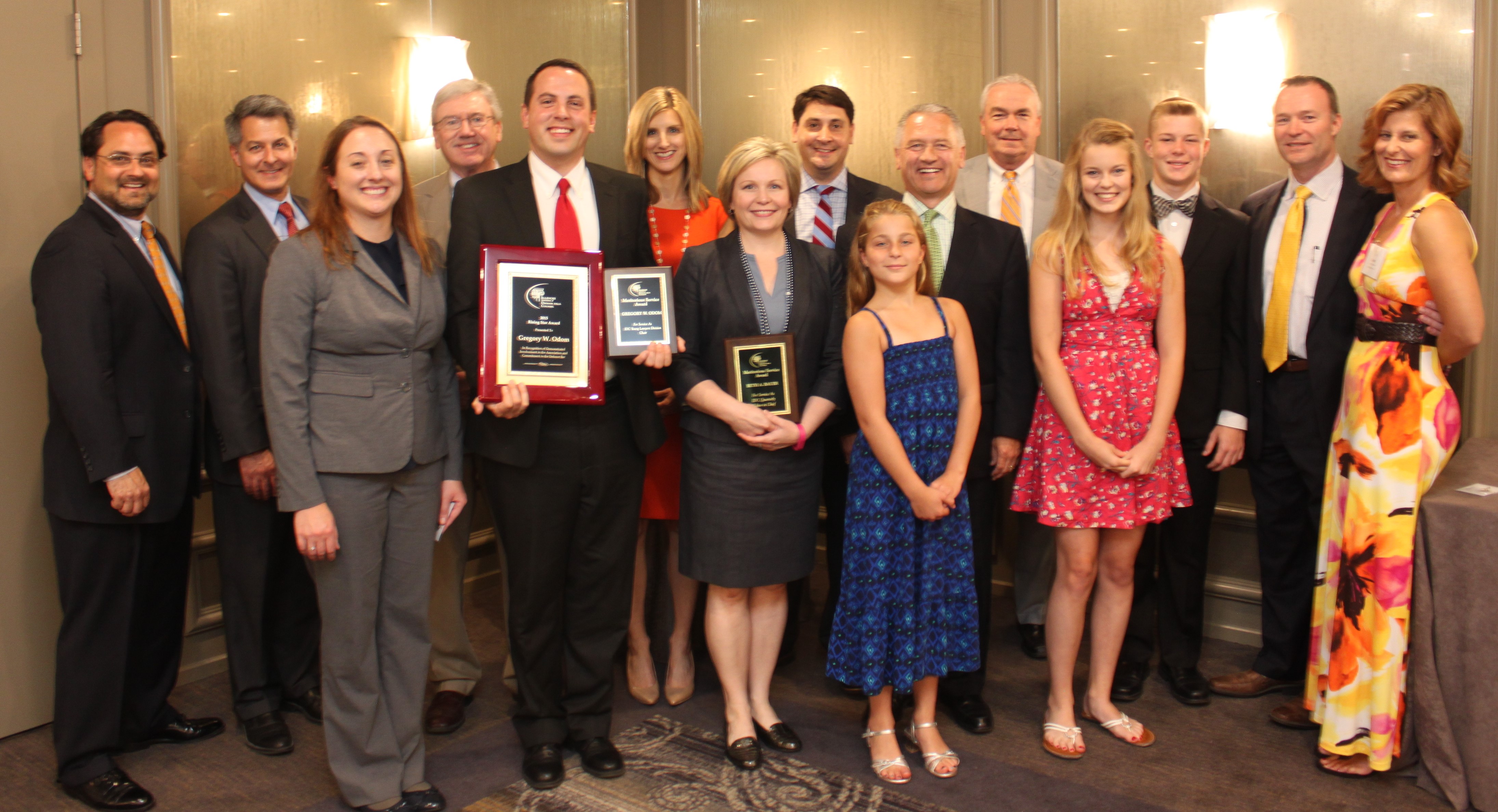 Group of 8 men (all wearing suits), 4 women (1 wearing a pantsuit and 3 wearing a dress), 2 female children (both wearing dresses), and one male child (wearing a tuxedo) standing in front of a ballroom wall. In the front row, HeplerBroom attorney Greg Odom is holding 2 award plaques, and to his left, HeplerBroom attorney Beth Bauer is holding 1 award plaque.