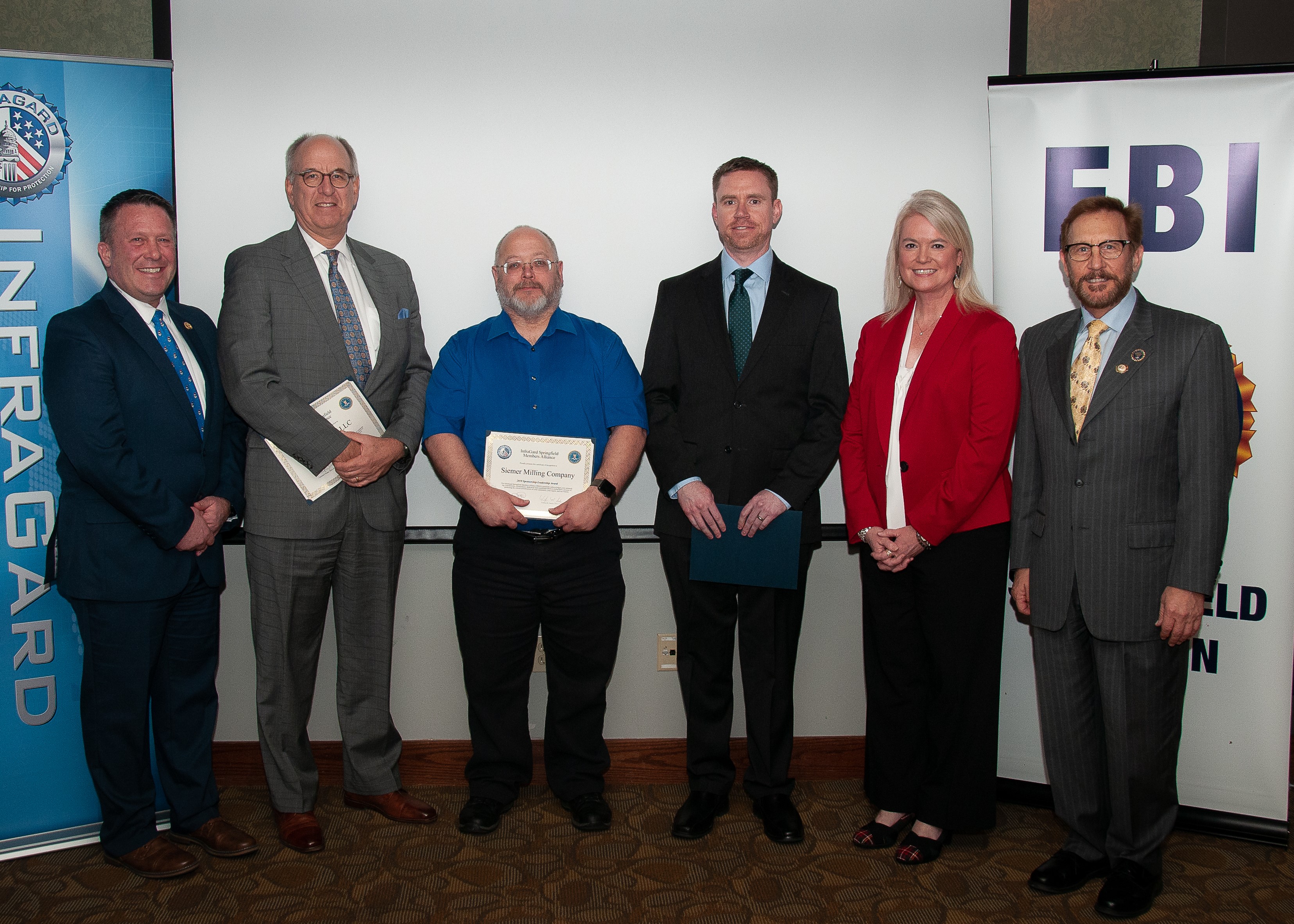 Group of 5 men and 1 woman standing. Two men are holding certificates. A vertical banner on the left of the group has "Infragard" printed down the side; at the top there is a portion of an Infragard logo visible. A vertical banner to the right of the group has "FBI" visible at the top; the rest of the banner is obscured by a man standing in front of it.