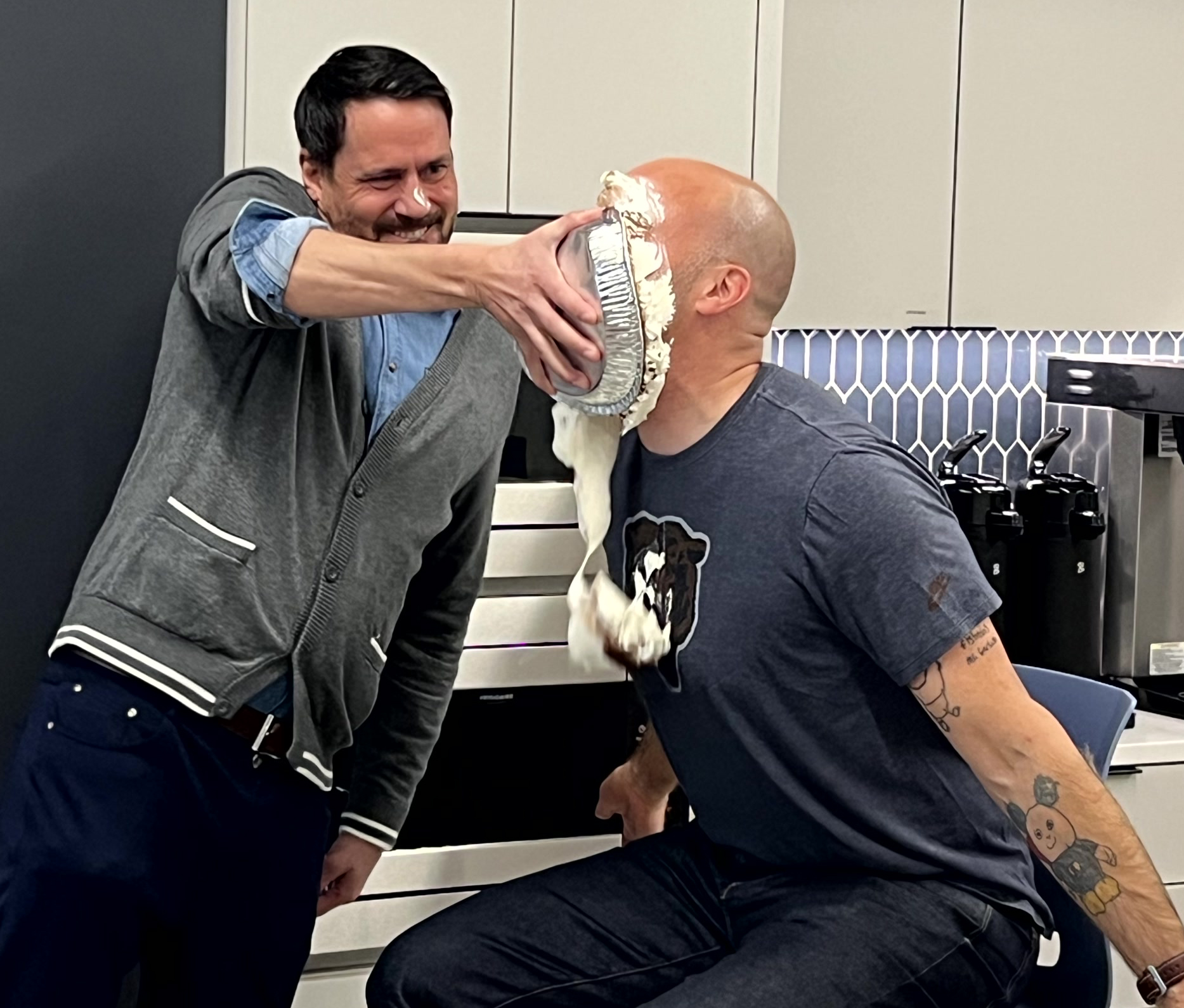 man on left shoving cream pie in face of man on right